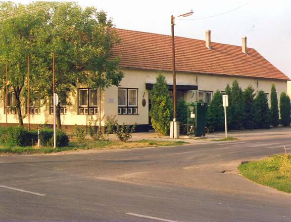 Das Bild zeigt ein eingeschossiges, längliches Gebäude mit einem Ziegeldach und mehreren Fenstern, umgeben von Bäumen und Büschen, an einer Straße mit einem Straßenlaternenpfahl und einem Transformator.