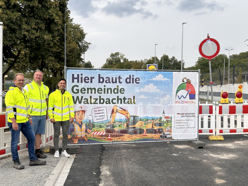 Drei Männer in gelben Warnwesten stehen neben einem Bauzaun mit einem Banner, auf dem "Hier baut die Gemeinde Walzbachtal" steht, vor einer Baustelle.