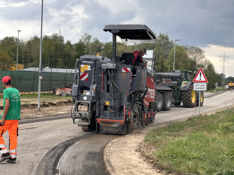 Eine Straßenbaumaschine ist auf einer Baustelle im Einsatz, während ein Arbeiter daneben steht und der Himmel bewölkt ist.