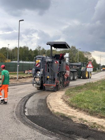 Eine Straßenbaumaschine ist auf einer Baustelle im Einsatz, während ein Arbeiter daneben steht und der Himmel bewölkt ist.