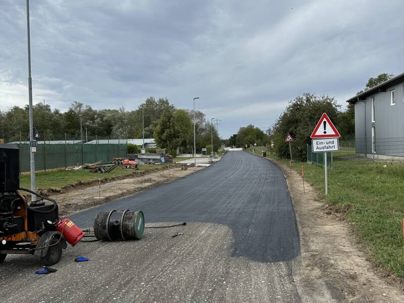Eine gerade Straße wird asphaltiert, mit Baugerät am Straßenrand und einem Warnschild über eine Ein- und Ausfahrt.