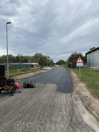 Eine gerade Straße wird asphaltiert, mit Baugerät am Straßenrand und einem Warnschild über eine Ein- und Ausfahrt.