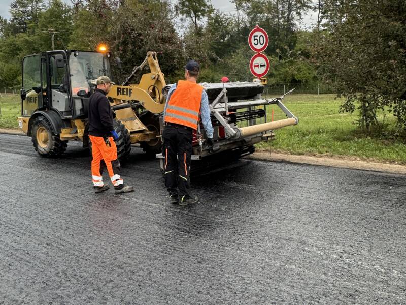 Zwei Arbeiter in orangefarbener Schutzkleidung stehen neben einer Straßenbaumaschine auf frisch asphaltiertem Straßenbelag, mit Verkehrsschildern im Hintergrund.