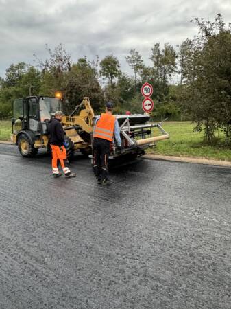 Zwei Arbeiter in orangefarbener Schutzkleidung stehen neben einer Straßenbaumaschine auf frisch asphaltiertem Straßenbelag, mit Verkehrsschildern im Hintergrund.