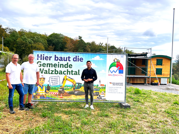 Drei Männer stehen vor einem Bauschild mit der Aufschrift "Hier baut die Gemeinde Walzbachtal" auf einer Wiese, im Hintergrund ist ein kleines Holzgebäude und Wald zu sehen.
