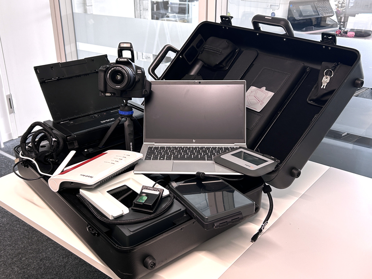 Ein Koffer, der auf einem Tisch geöffnet ist, enthält verschiedene technische Geräte wie eine Kamera, einen Laptop, einen Router, ein Kabel und weitere elektronische Zubehörteile.