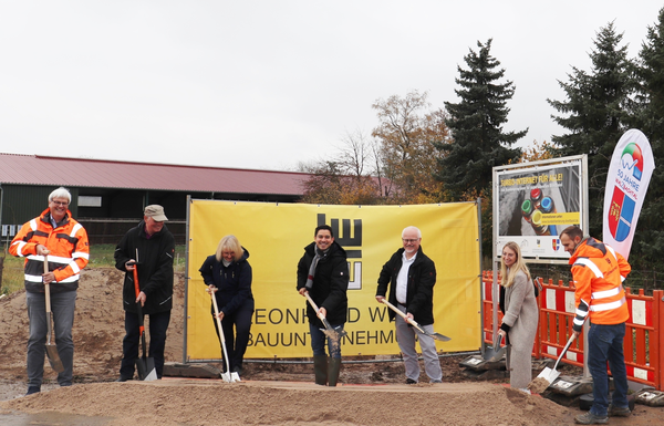 Eine Gruppe von Menschen in Arbeitskleidung hält Schaufeln und steht vor einem gelben Banner bei einer Baustelle.