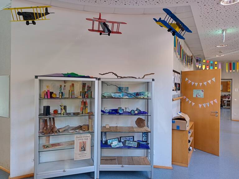 In einem Raum stehen zwei Vitrinen mit bunt gestalteten Objekten, fliegenden Modellflugzeugen an der Decke und einer dekorierten Holztür rechts.