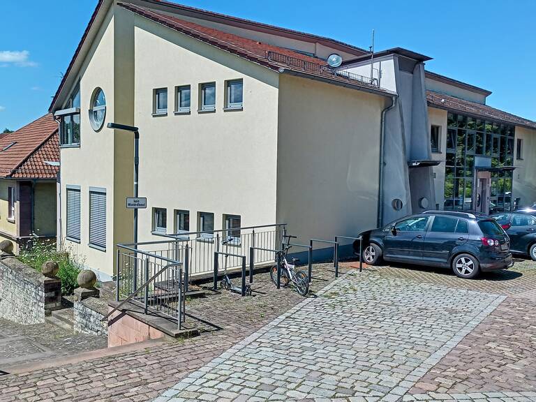 Ein modernes Gebäude mit einem schrägen Dach und mehreren Fenstern hat vor seiner Fassade eine Rampe mit Geländer, daneben parken Autos und ein Fahrrad steht an der Rampe.