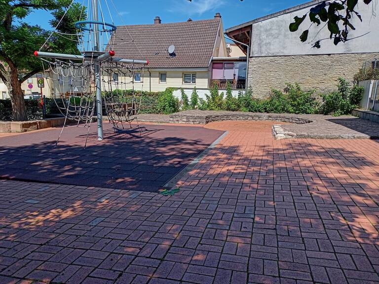 Ein leerer Spielplatz mit einem Klettergerüst aus Seilen steht auf einem gepflasterten Boden, umgeben von Büschen und Häusern im Hintergrund.
