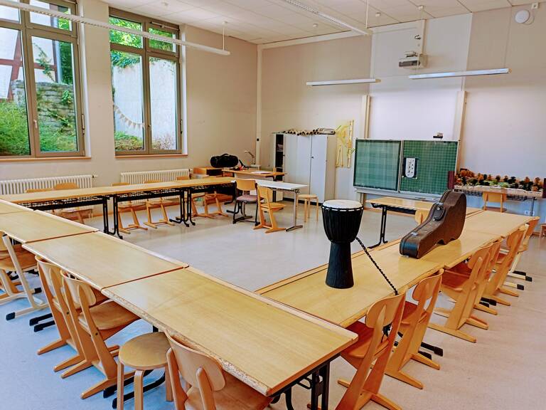 Ein Klassenraum mit U-förmig angeordneten Tischen, auf denen eine Djembe-Trommel und ein Gitarrenkoffer stehen, sowie eine Tafel und Fenster mit Blick auf eine grüne Außenanlage.