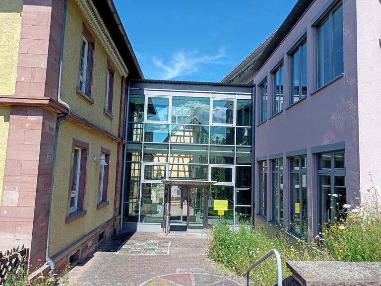 Ein modernes Gebäude mit Glasfront verbindet zwei ältere Gebäude, eins mit gelber und das andere mit grauer Fassade, unter einem blauen Himmel.