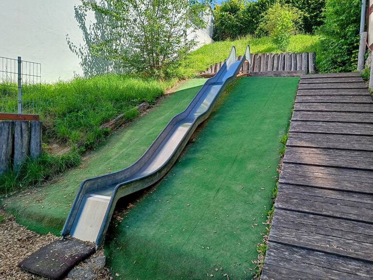 Das Bild zeigt eine Rutsche aus Metall, die in eine grasbewachsene Fläche eingebettet ist, neben einem Holzweg hinaufführt.