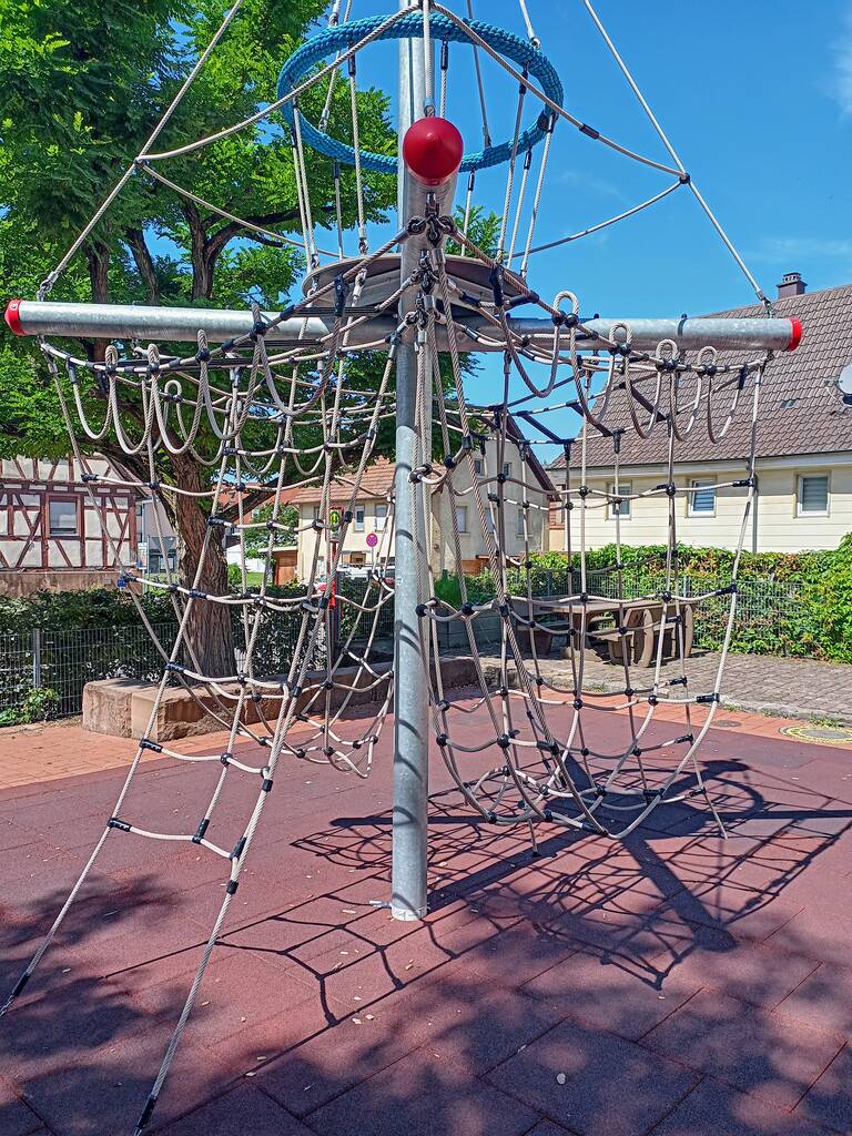 Ein Klettergerüst aus Seilen und Metallstangen steht auf einem Spielplatz mit weichem Bodenbelag, umgeben von Bäumen und Wohnhäusern.