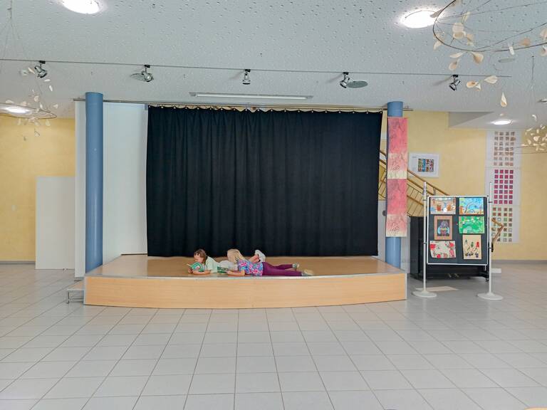 Zwei Kinder liegen auf einer kleinen Bühne vor einem geschlossenen schwarzen Vorhang und lesen in einem hellen Raum mit Kunstausstellungen an der Wand und auf Stelltafeln.