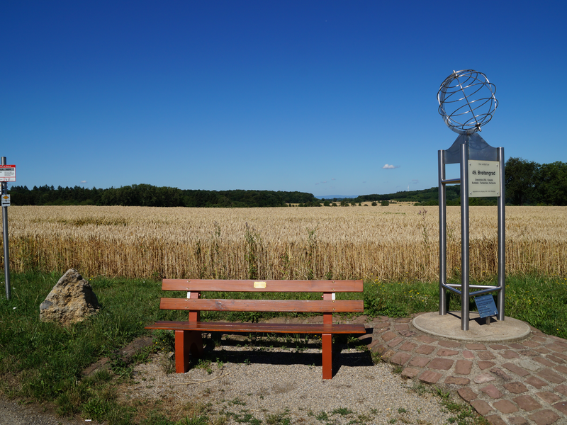 Eine Holzbank steht vor einem Kornfeld, begleitet von einem Metallschild und einer Skulptur, bei klarem Himmel.