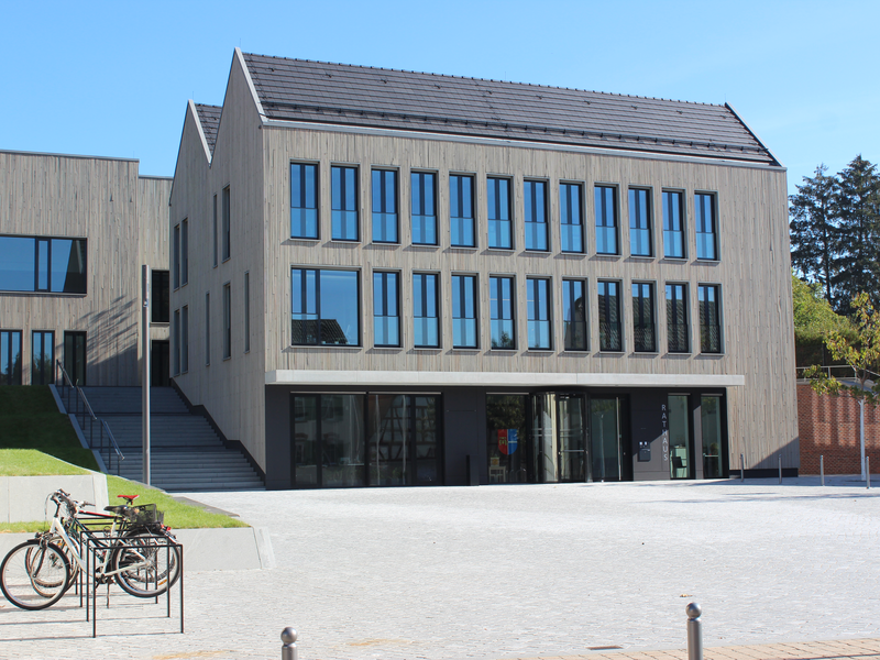 Ein modernes, dreistöckiges Gebäude mit vielen großen, rechteckigen Fenstern steht in einer gepflasterten Umgebung, neben der einige Fahrräder geparkt sind.