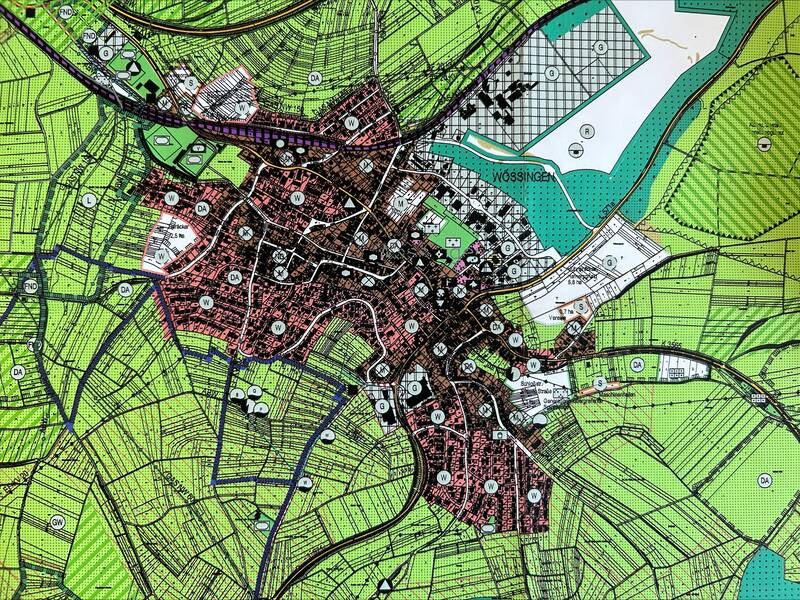 Der Stadtplan zeigt eine detaillierte Darstellung eines städtischen Gebiets, umgeben von grünen landwirtschaftlichen Flächen, mit Straßen, Gebäuden und einem Gewässer am oberen rechten Rand.