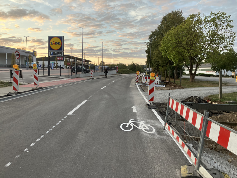 Die Aufnahme zeigt eine Straße bei Sonnenuntergang mit Fahrbahnmarkierungen für Radfahrer und Baustellenabsperrungen sowie ein nahegelegenes Lidl-Geschäft.
