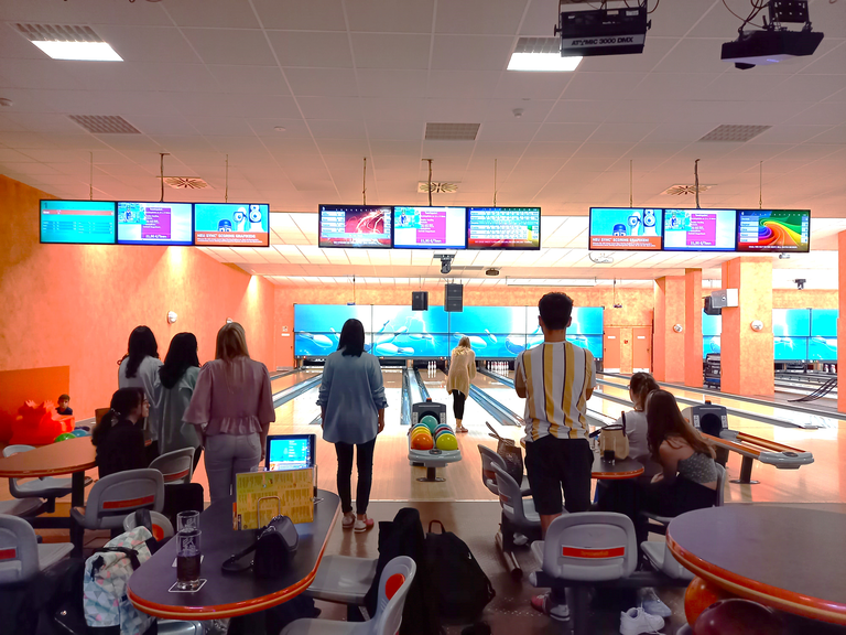 Eine Gruppe von Personen steht und sitzt vor Bowlingbahnen in einer Halle, während sie einem Spiel zuschauen.