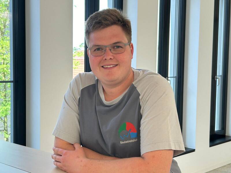 Herr Florian Schwarz, ein lächelnder Mann mit Brille und einem "Baubetriebshof" T-Shirt sitzt an einem Tisch vor großen Fenstern.