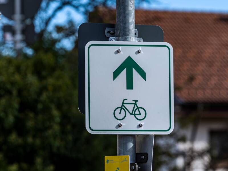 Ein weißes Schild mit einem grünen Fahrrad und Richtungspfeil als Wegweiser für einen Radweg. Das Schild ist an einem Laternenpfosten befestigt.