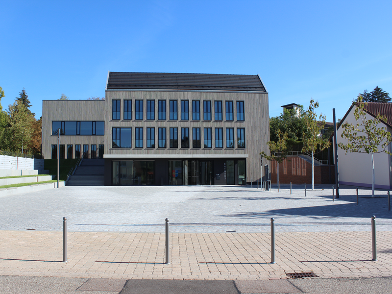Das Bild zeigt ein modernes Gebäude mit mehreren großen Fenstern und einem flachen Dach, das von einem gepflasterten Platz umgeben ist, auf dem einige Bäume stehen.