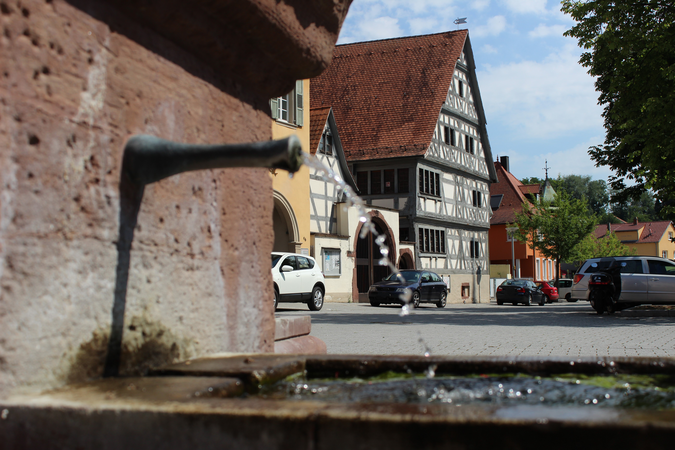 Ein Brunnen mit fließendem Wasser im Vordergrund und einem Fachwerkhaus sowie parkenden Autos im Hintergrund.