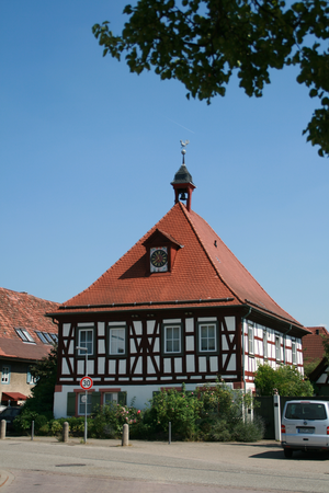 Ein traditionelles Fachwerkhaus mit rotem Dach, Uhrtürmchen und Wetterfahne steht an einer Straße neben geparkten Fahrzeugen an einem sonnigen Tag.