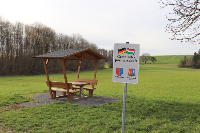 Überdachter Picknickplatz auf grüner Wiese. Davor ein Schild mit dem Wappen von Walzbachtal und Bacsbokod