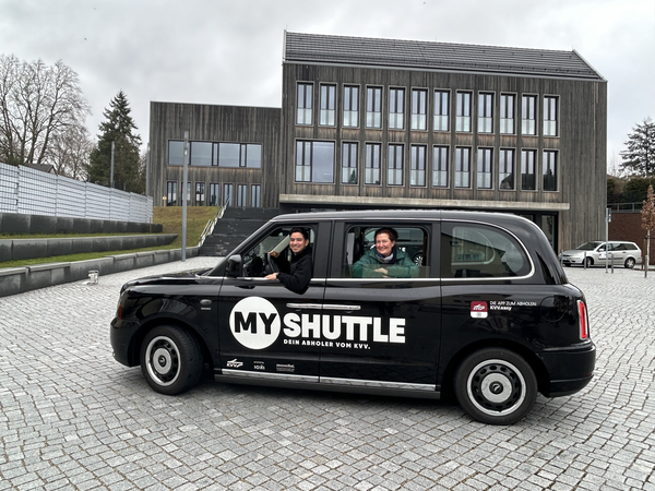 Das Bild zeigt ein schwarzes Auto mit dem Aufdruck "MY SHUTTLE", in dem zwei Personen sitzen, vor einem modernen Gebäude mit großen Fenstern.