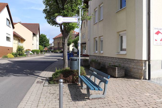 Mitfahrbank mit Hinweisschild neben der Wössinger Str.. Rechts und Links Häuser