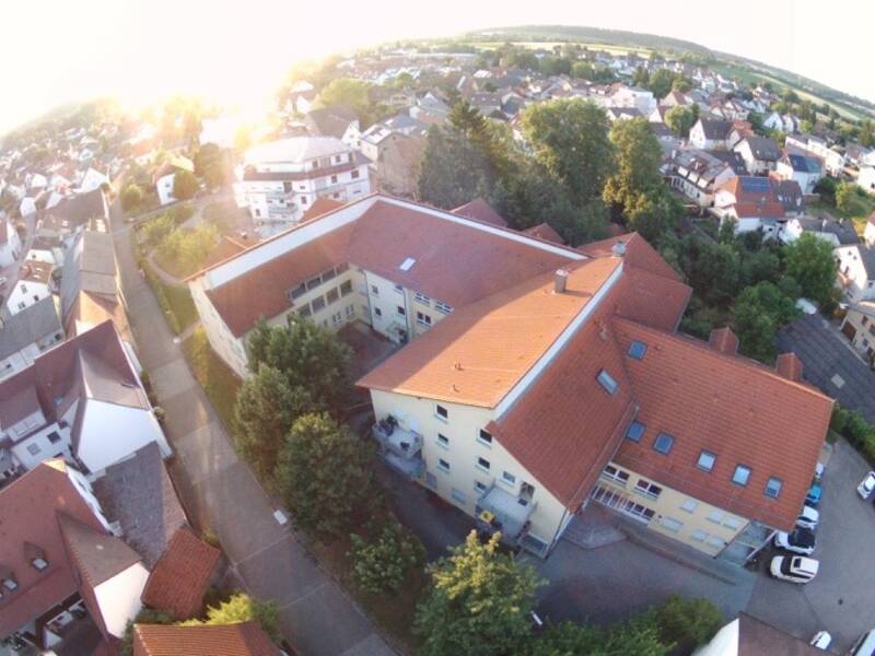 Das Bild zeigt eine luftige Perspektive eines Gebäudes mit roten Ziegeldächern, Bäumen und Straßen in heller Abendsonne.