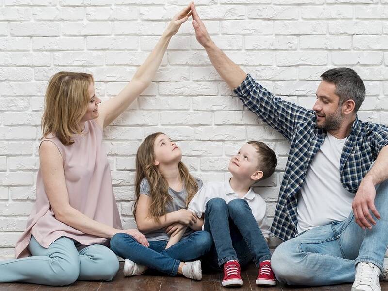 Eine Familie sitzt auf dem Boden vor einer weißen Ziegelwand; die Eltern halten ihre Hände über den beiden Kindern wie ein Dach zusammen.